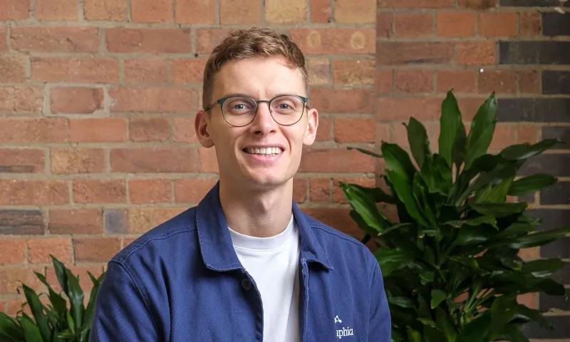 A member of the Biographica team, smiling, in a navy Biographica jacket in front of a brick wall and green plants